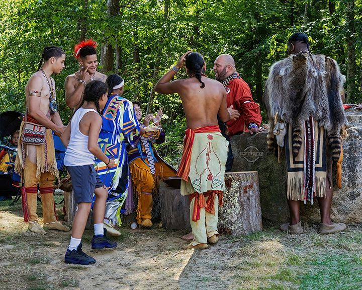 Group of people in Native American attire gathered outdoors, preparing or sharing something, possibly a ceremony.