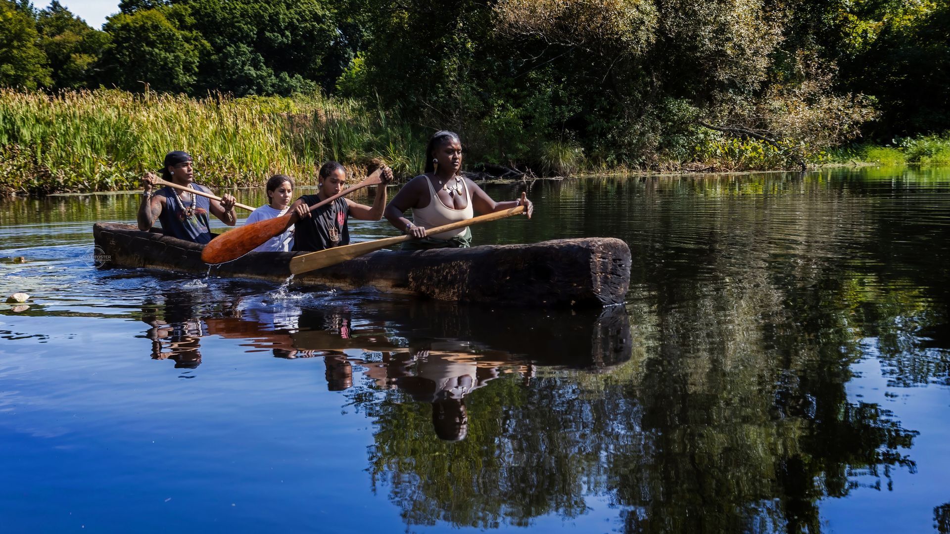 People paddling a log canoe on a calm lake, surrounded by greenery.