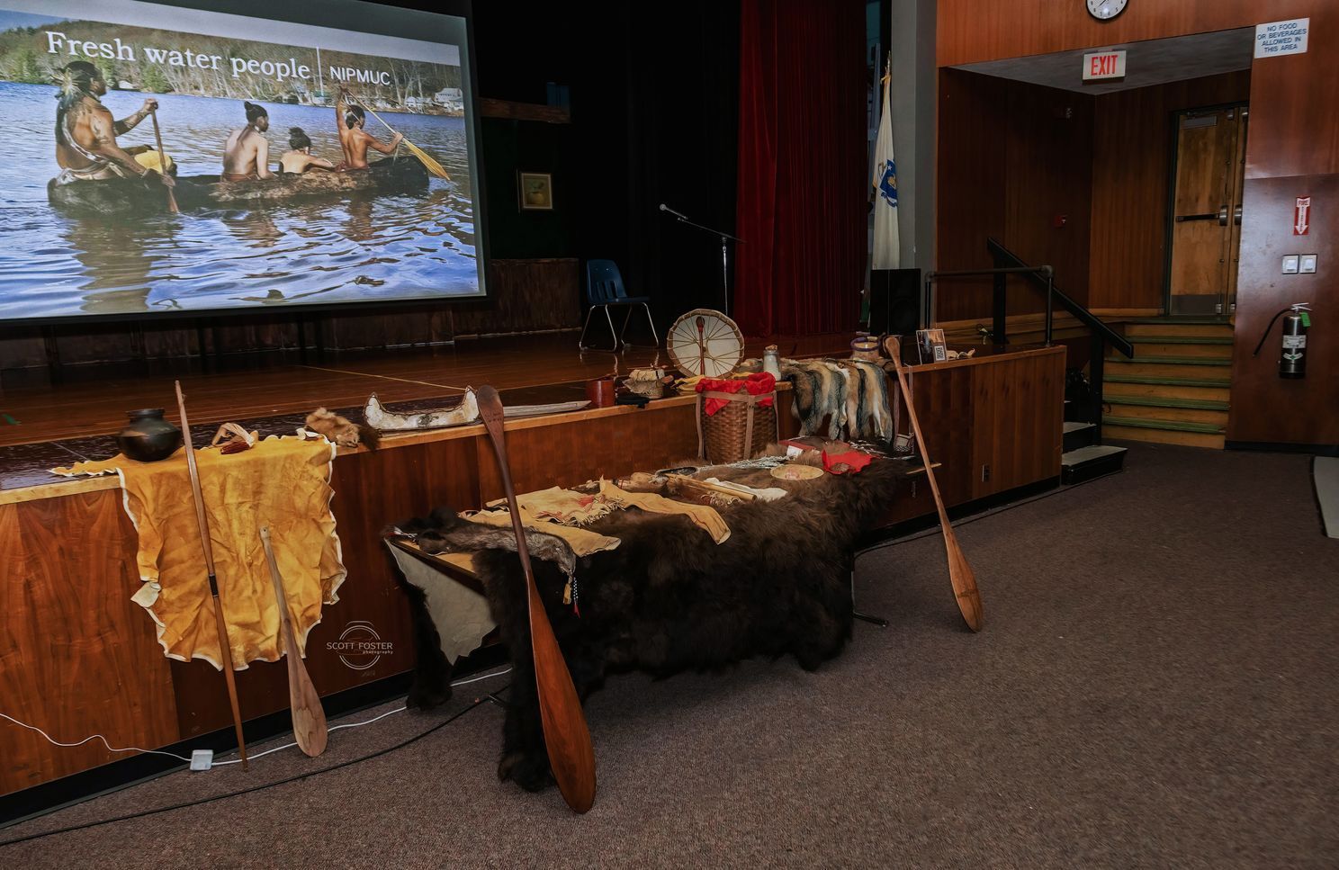 Display of Indigenous artifacts: paddles, pelts, dried fish, on a stage with a projected image of canoes on water.