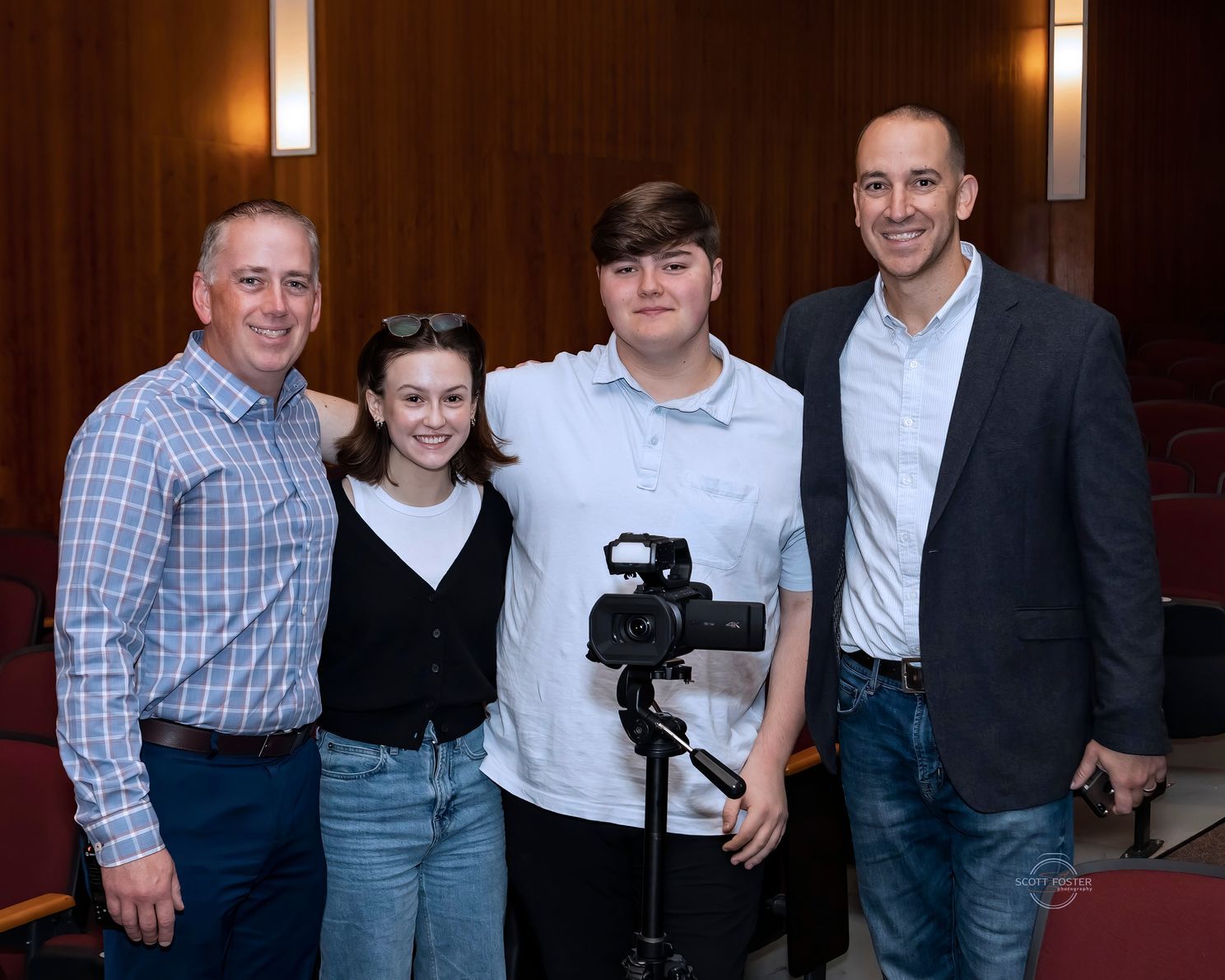 Four people pose for a photo in a theater. Two men flank two younger people, all smiling. A camera sits on a tripod.