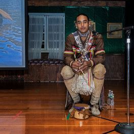 Person in traditional clothing sits, holding object, with a drum, microphone, and water bottle on a stage.