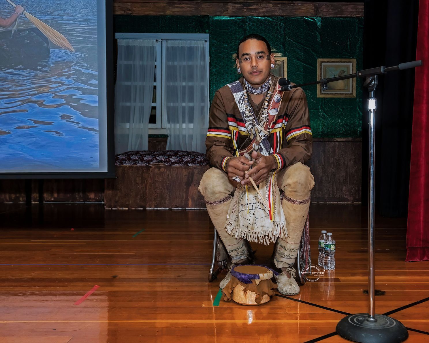Man in traditional Native American attire sits on stage, holding instruments.
