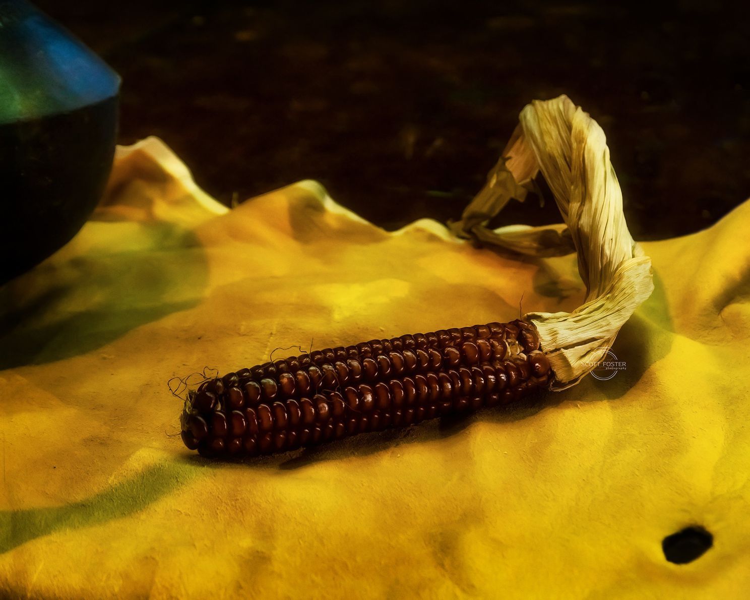 Ear of purple corn with husk on yellow cloth, near dark blue vase.