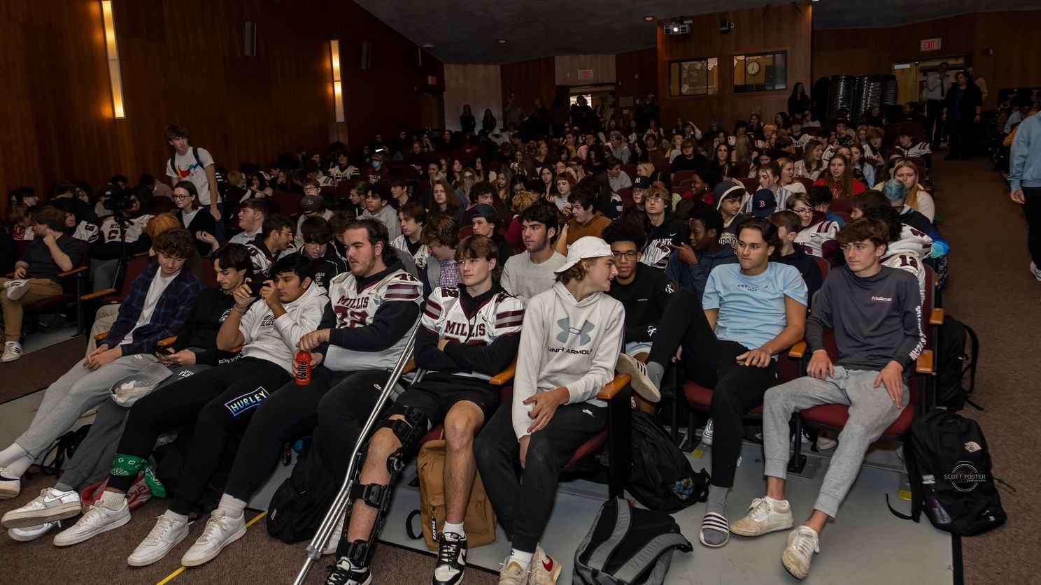 Audience in an auditorium, many wearing athletic attire, attentively facing forward.