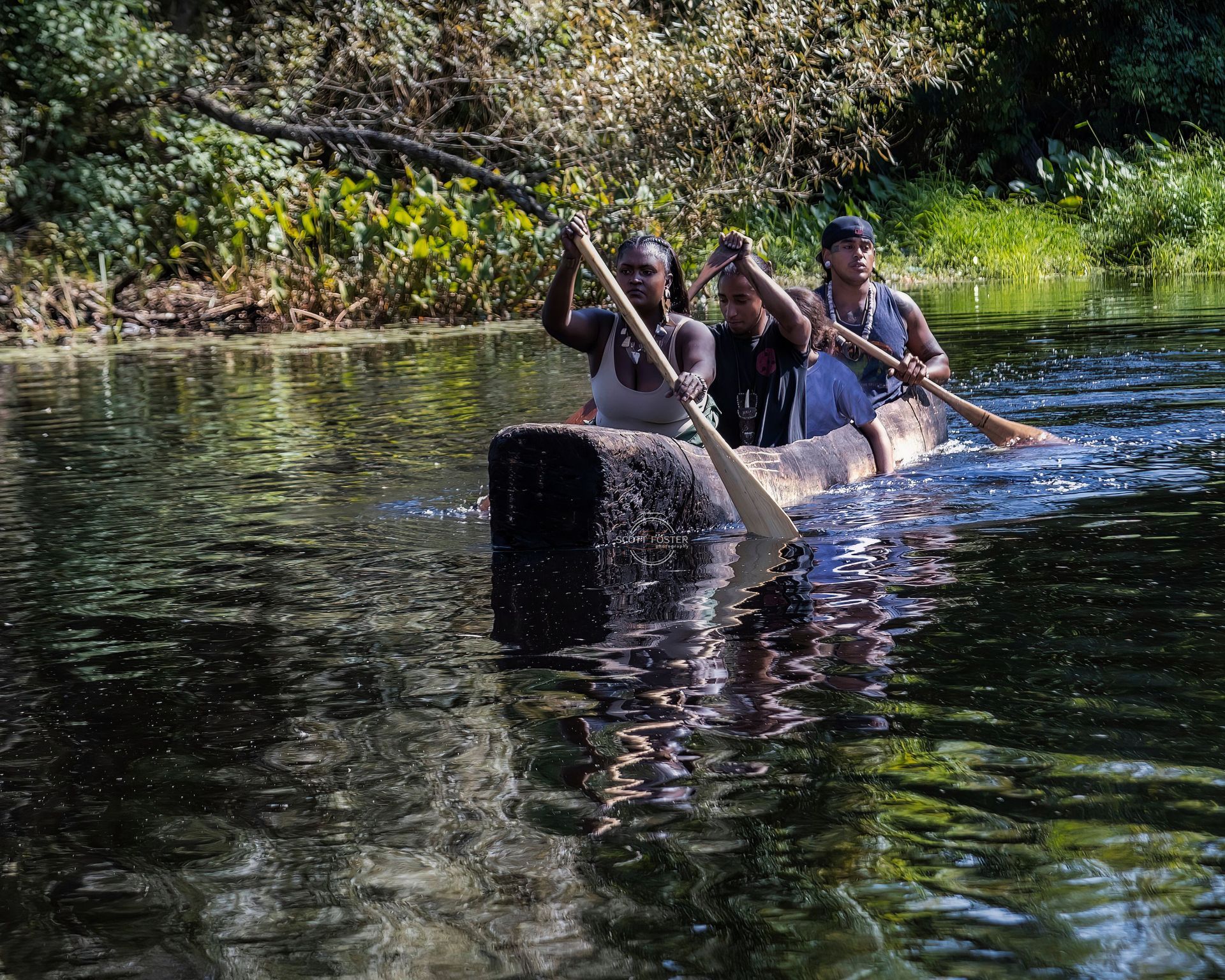 Three people paddling a dugout canoe on a dark river with lush green foliage in the background.