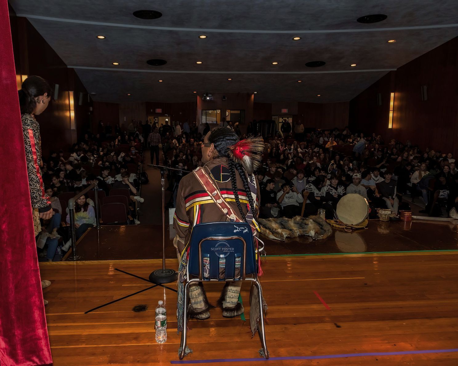 Person in Native American attire on stage; audience in auditorium.