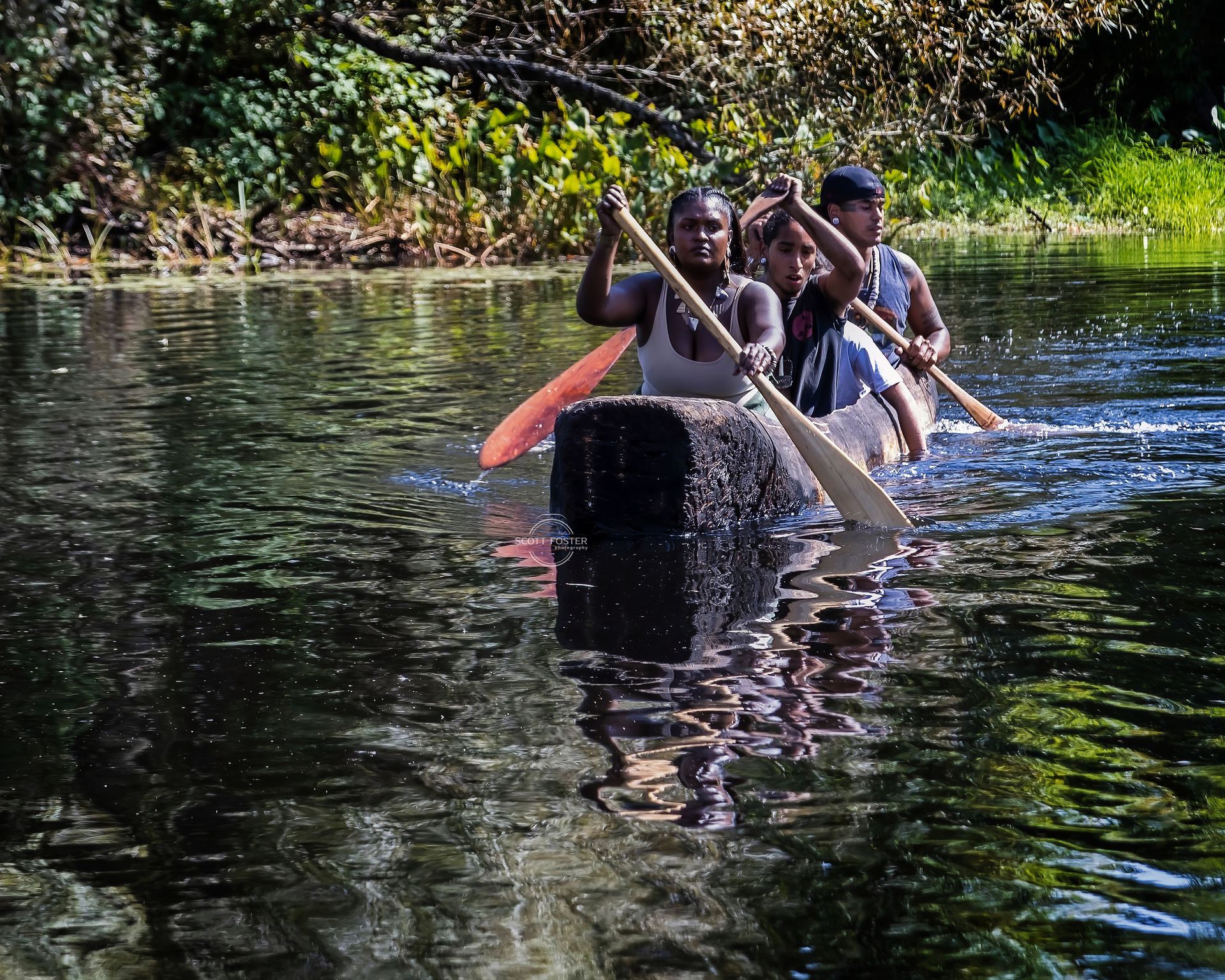 People paddling a canoe on a dark, reflective river surrounded by green foliage.