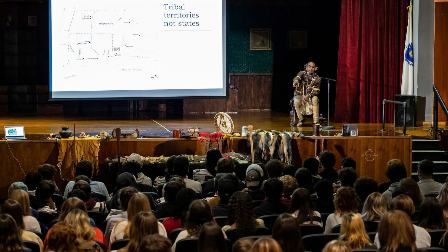 Audience seated, listening to a speaker on stage with a screen and display of artifacts.