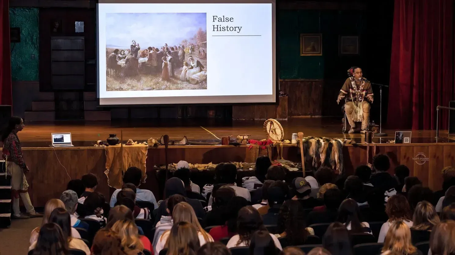 Presentation on stage with presenter, projector screen, and audience.