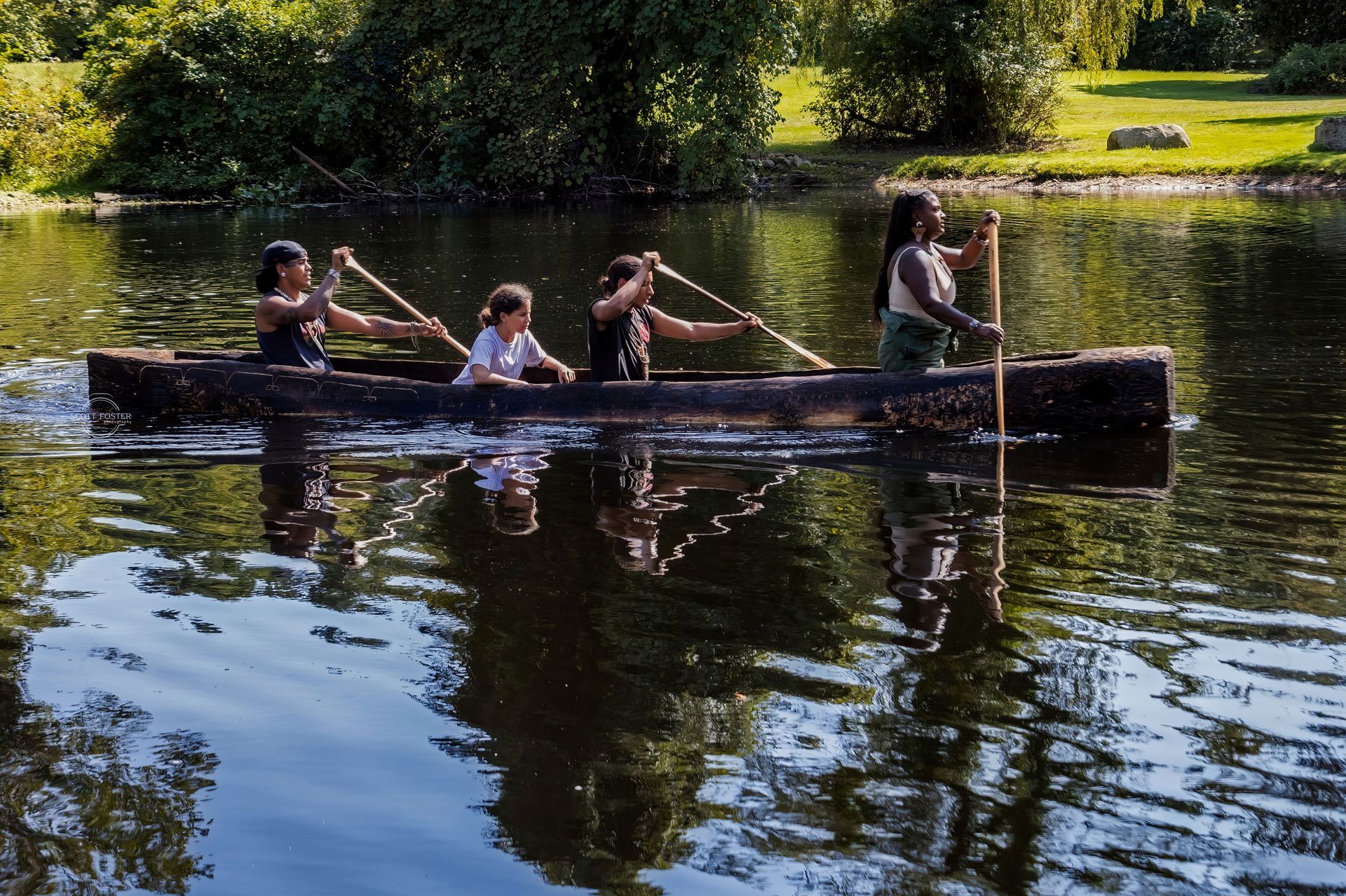 Four people paddling a long, dark canoe on a reflective body of water.