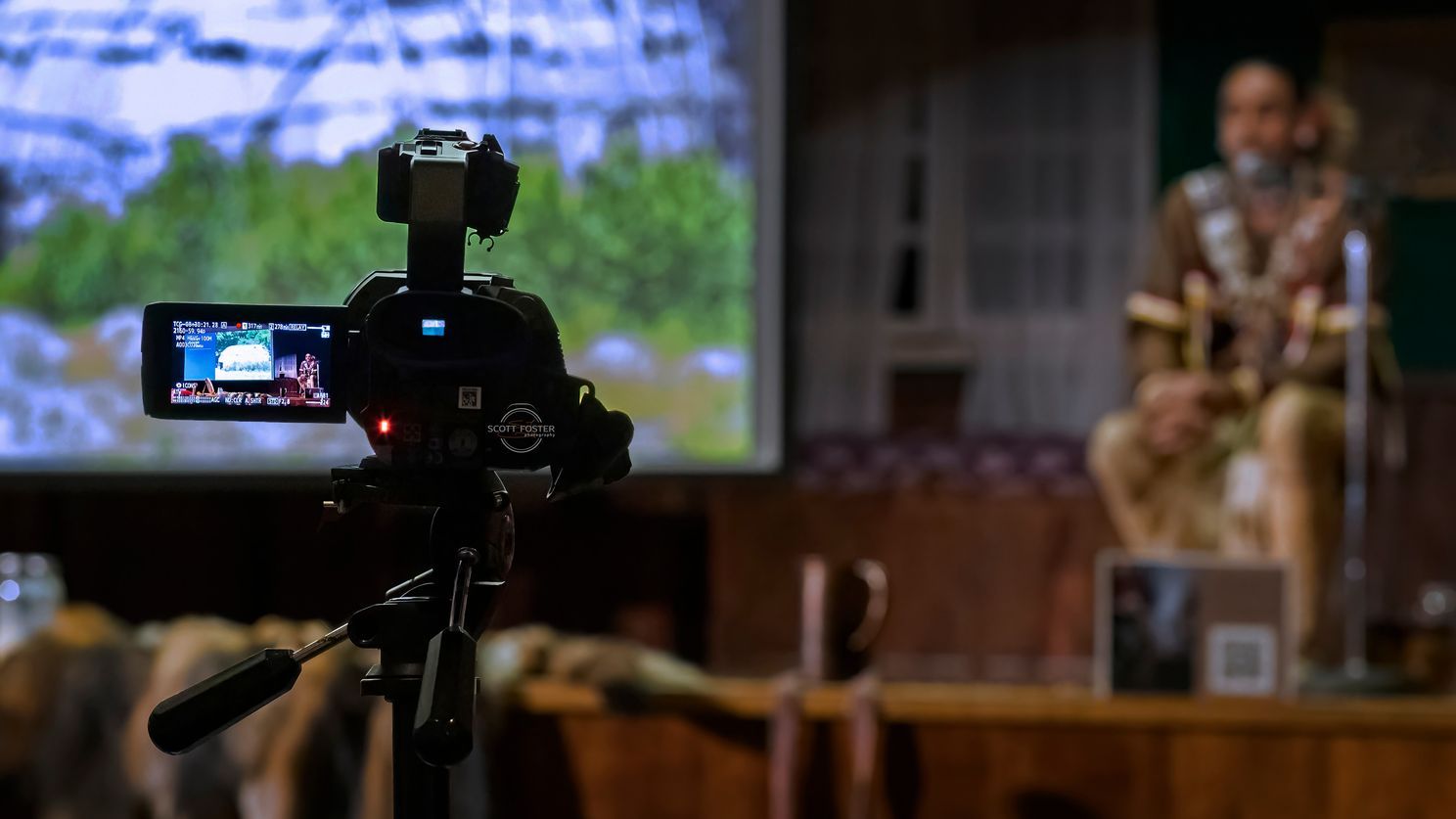 Camera filming a person on stage with a backdrop of a nature scene.