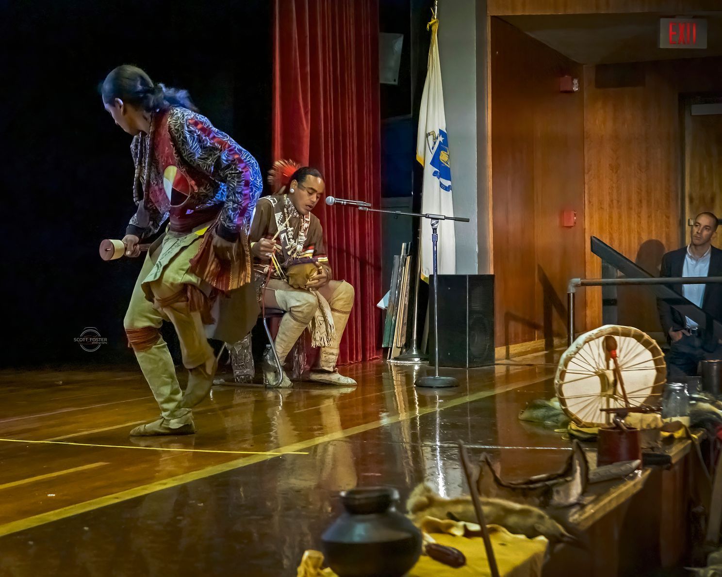 Two performers on stage in Native American attire; one dancing, the other drumming. Audience member watches.