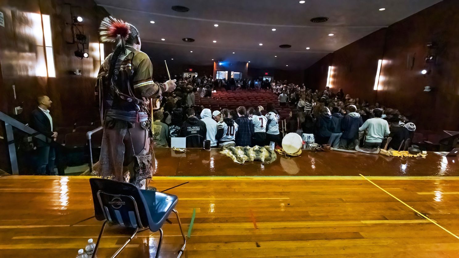 Indigenous person in regalia on stage speaking to large audience in auditorium.