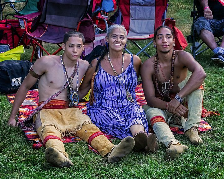 Three people seated on grass; a woman in a blue dress smiles between two individuals, both in traditional attire.