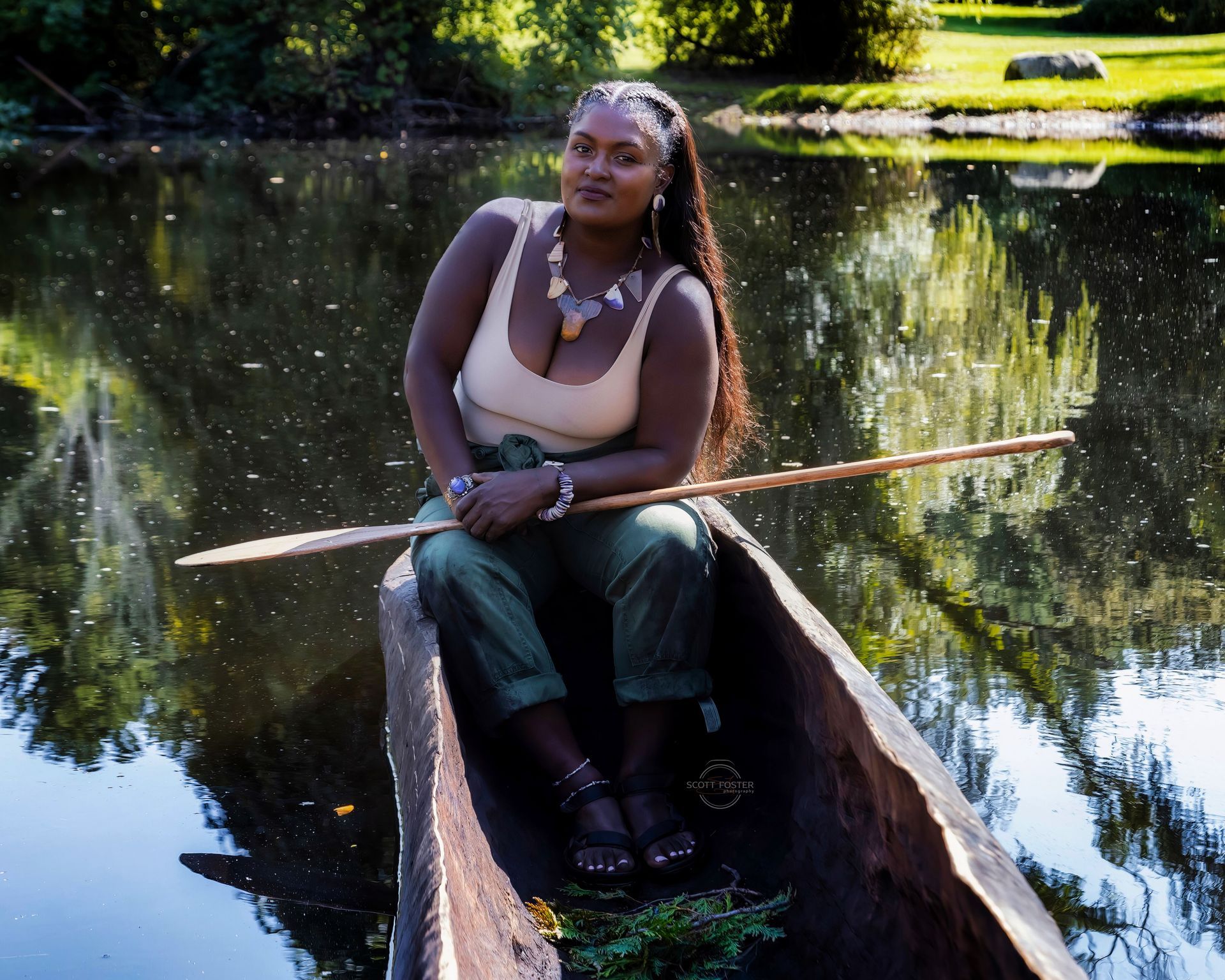 Woman in a wooden boat on a lake, holding a pole. She wears a cream top, green pants, jewelry.