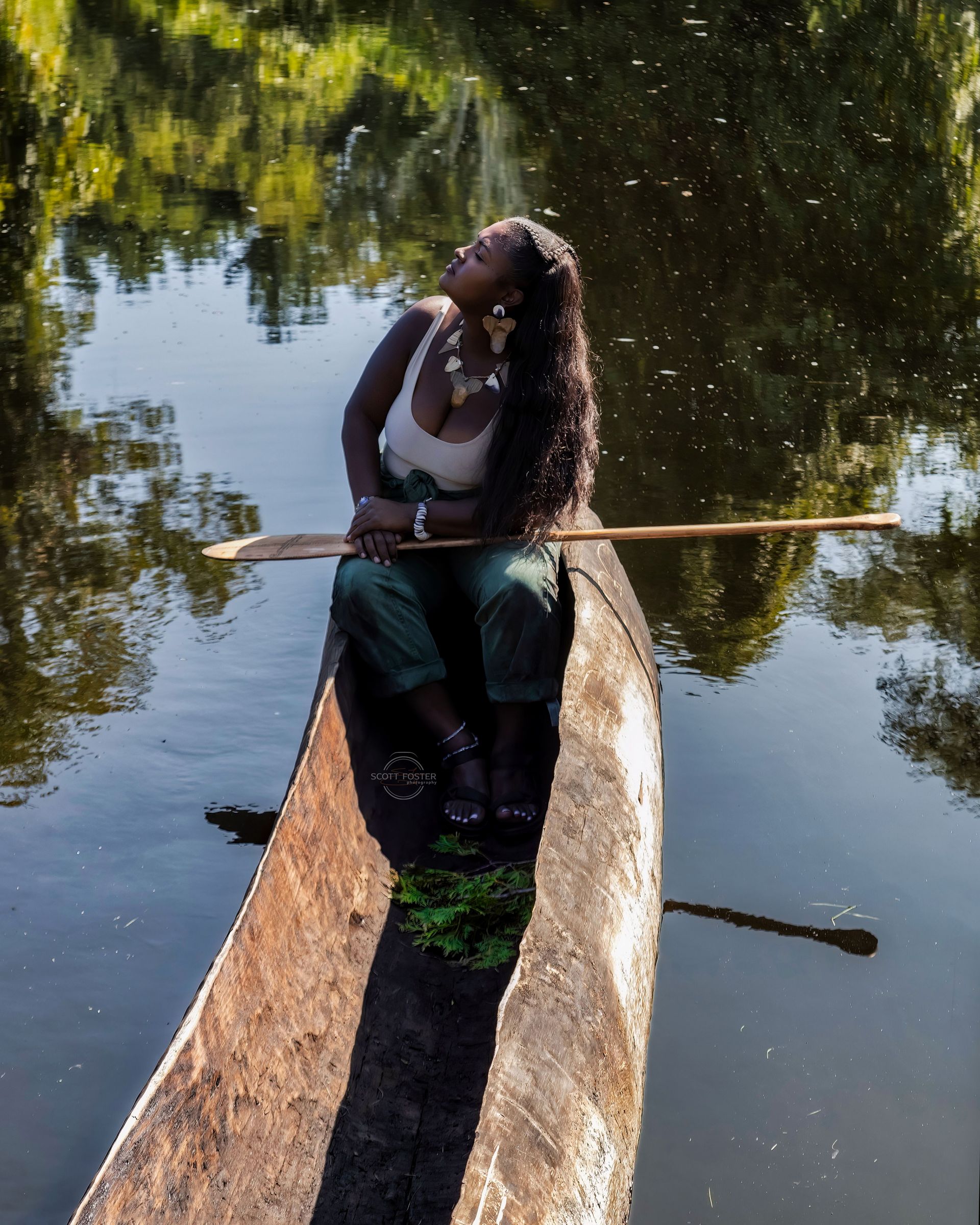 Woman in boat, looking up, holding paddle. Boat in water, trees reflected.