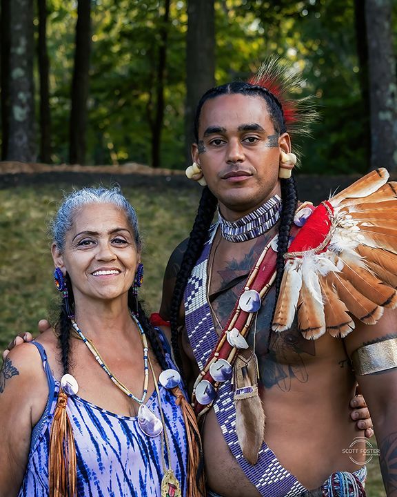 Woman and man in traditional Native American attire, smiling, standing outdoors. Man has headdress and chest covering.