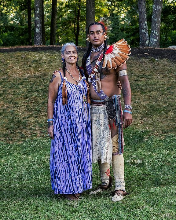 Woman in blue dress stands beside a person in Native American regalia on grassy hillside.