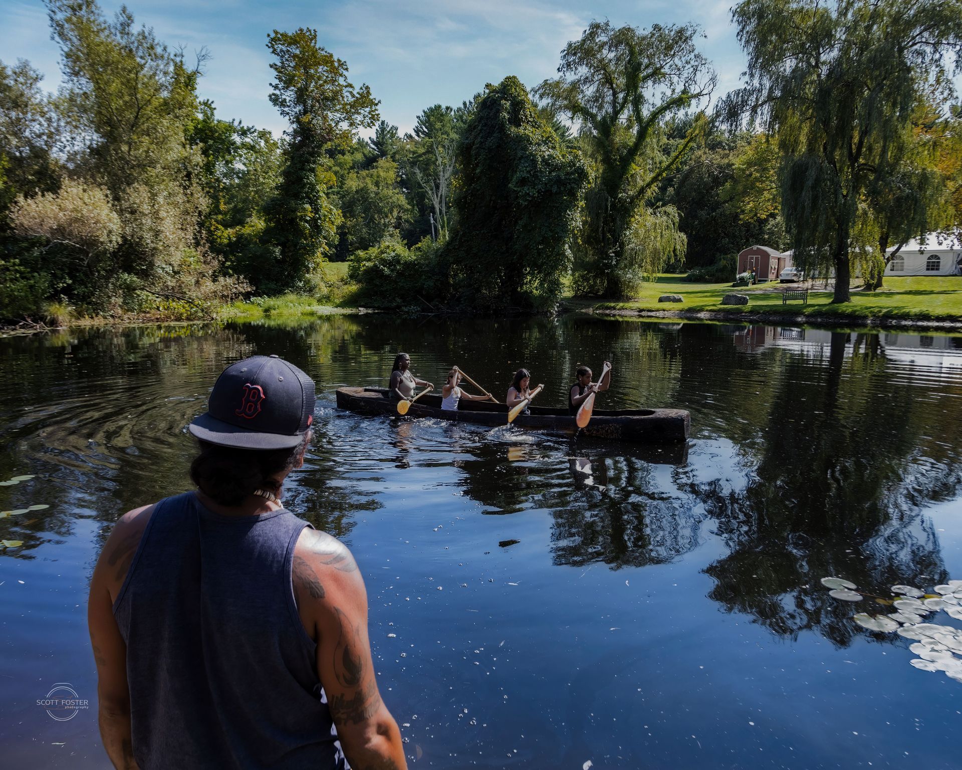 Man watches four people paddling a dark wooden vessel on calm water, surrounded by trees.