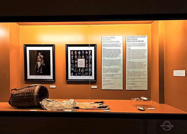 Museum display: framed photos, text panels, basket, fabric, and small objects on a brown surface, against an orange wall.