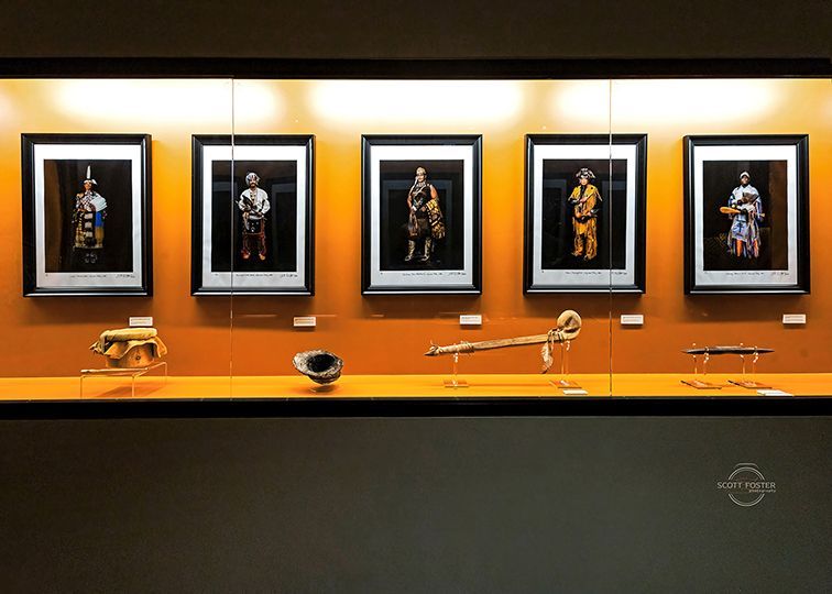 Museum display: five framed figures, various artifacts on a shelf below. Orange walls and a dark frame.