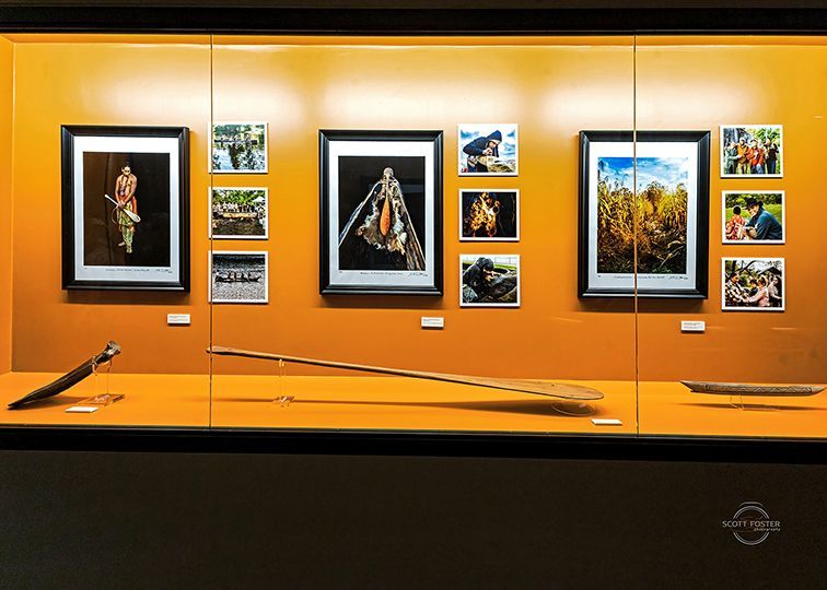 Museum display with framed photos and artifacts: long paddle, traditional attire. Photos show fields and people.