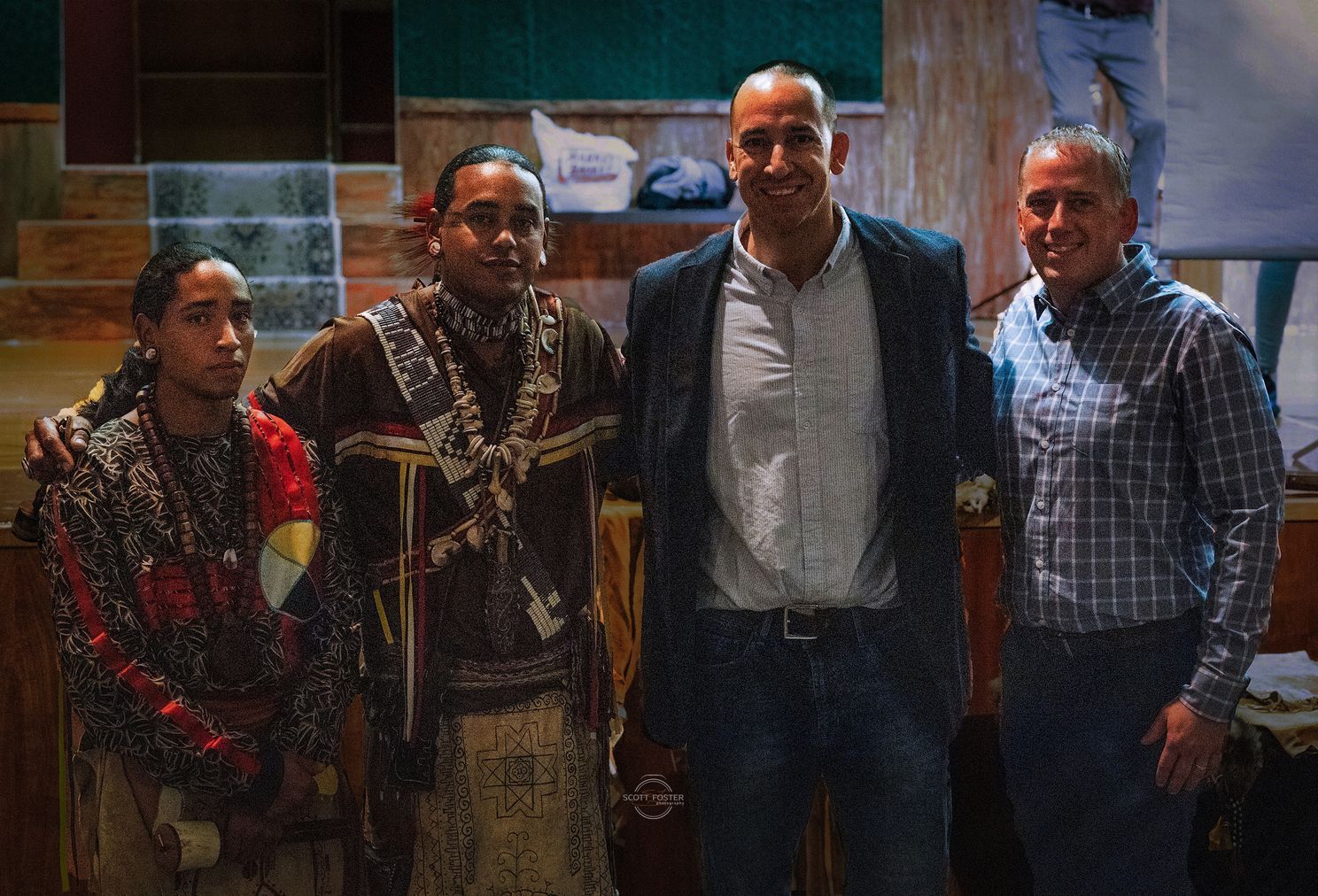 Four people standing together. Two in traditional Indigenous attire, two in business casual clothes.