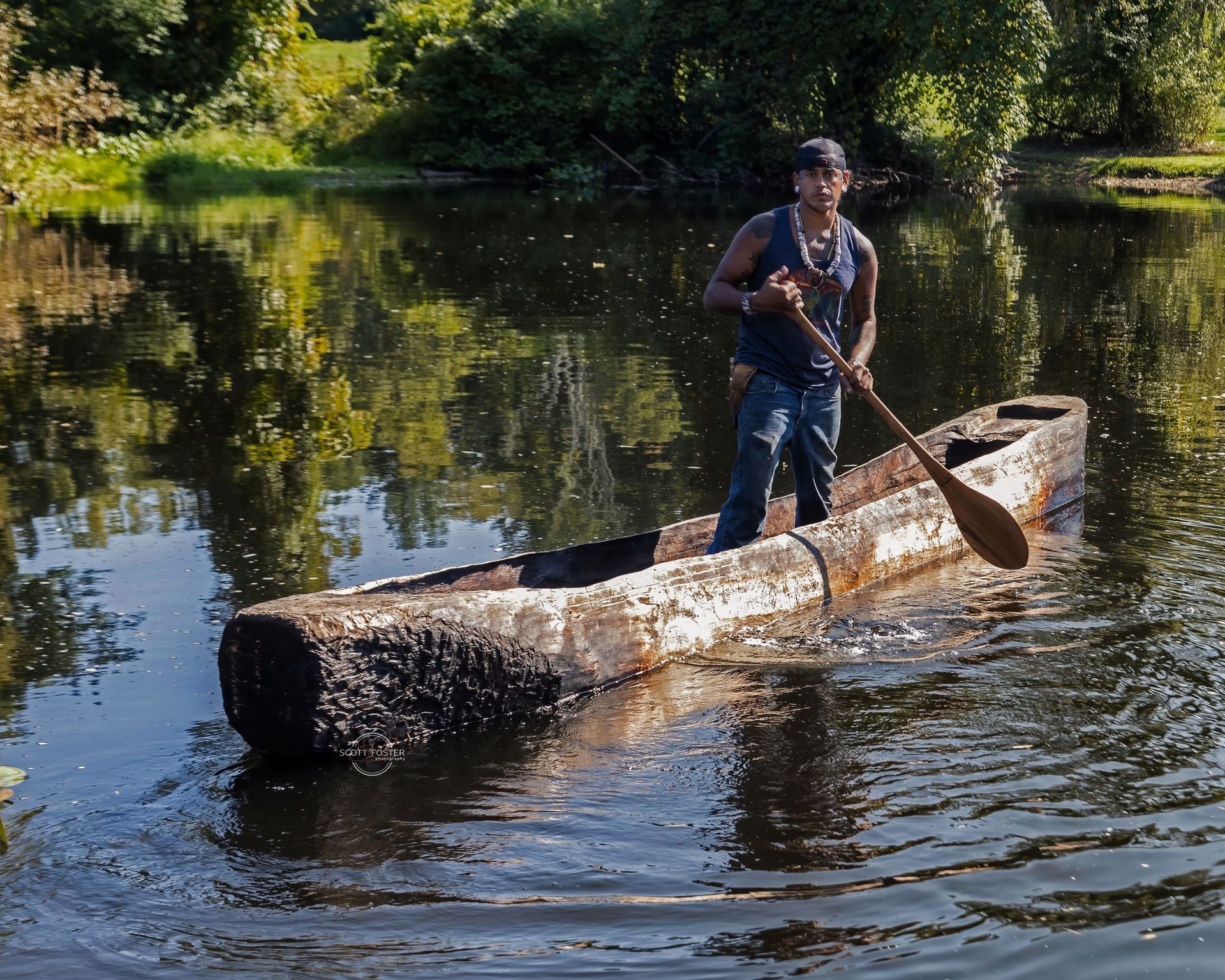Man paddles a wooden dugout canoe on a calm river. Green trees reflect in the water.