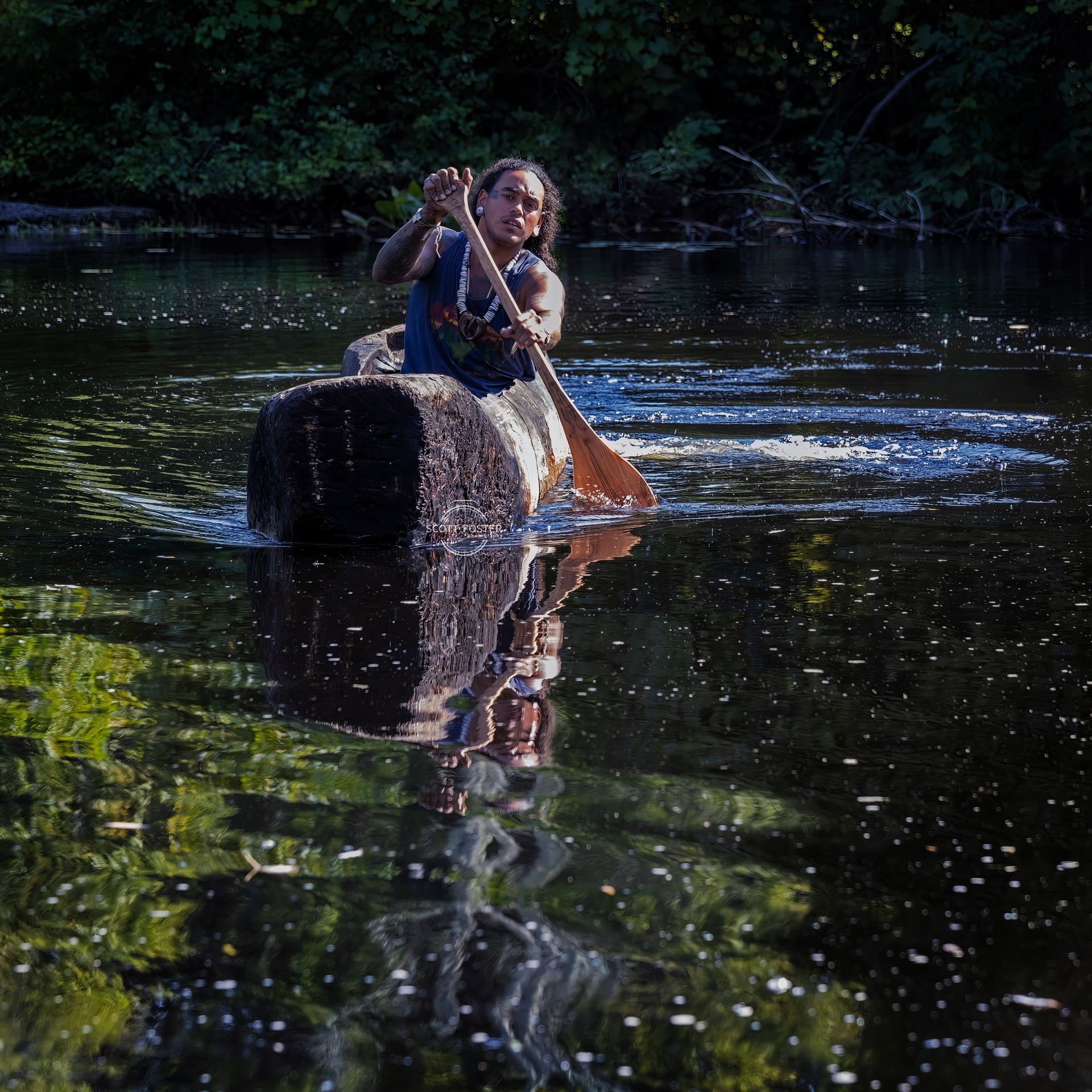 Person paddles a wooden canoe on a dark, reflective river with forest backdrop. Water ripples.