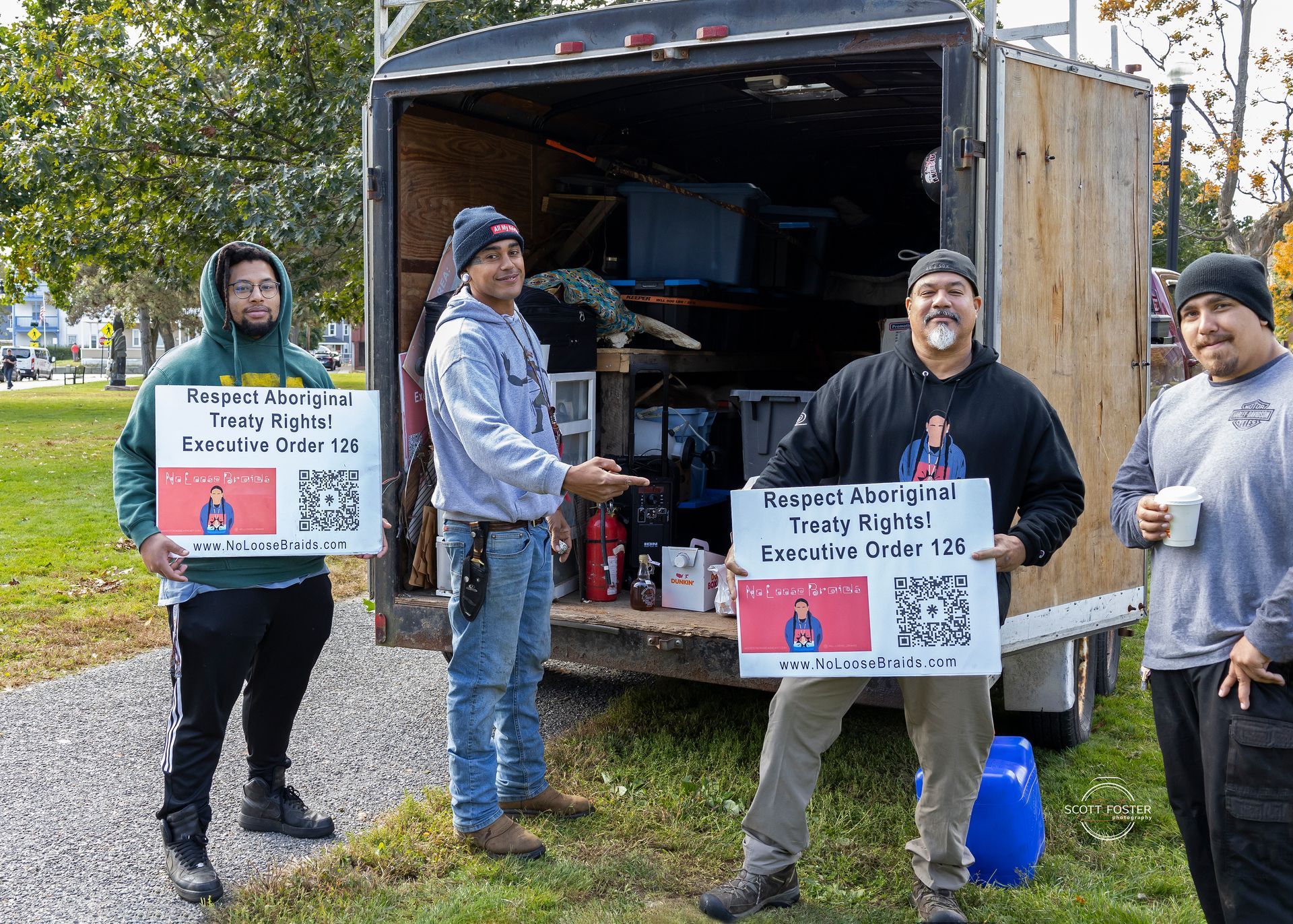 Four men holding signs in front of a trailer; the signs display text and a QR code.