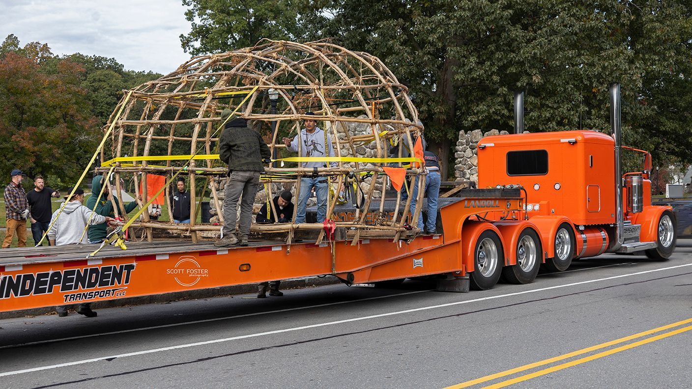 An orange semi-truck transports a large wooden dome structure. People stand on the trailer.