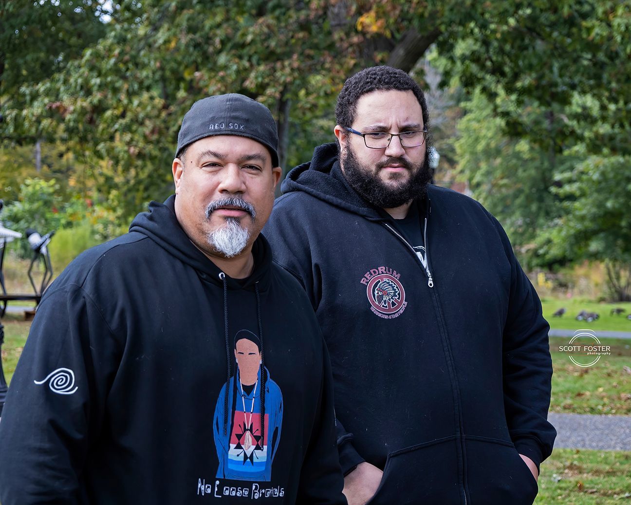 Two men in black hoodies stand outdoors, looking at the camera. Trees and foliage are in the background.