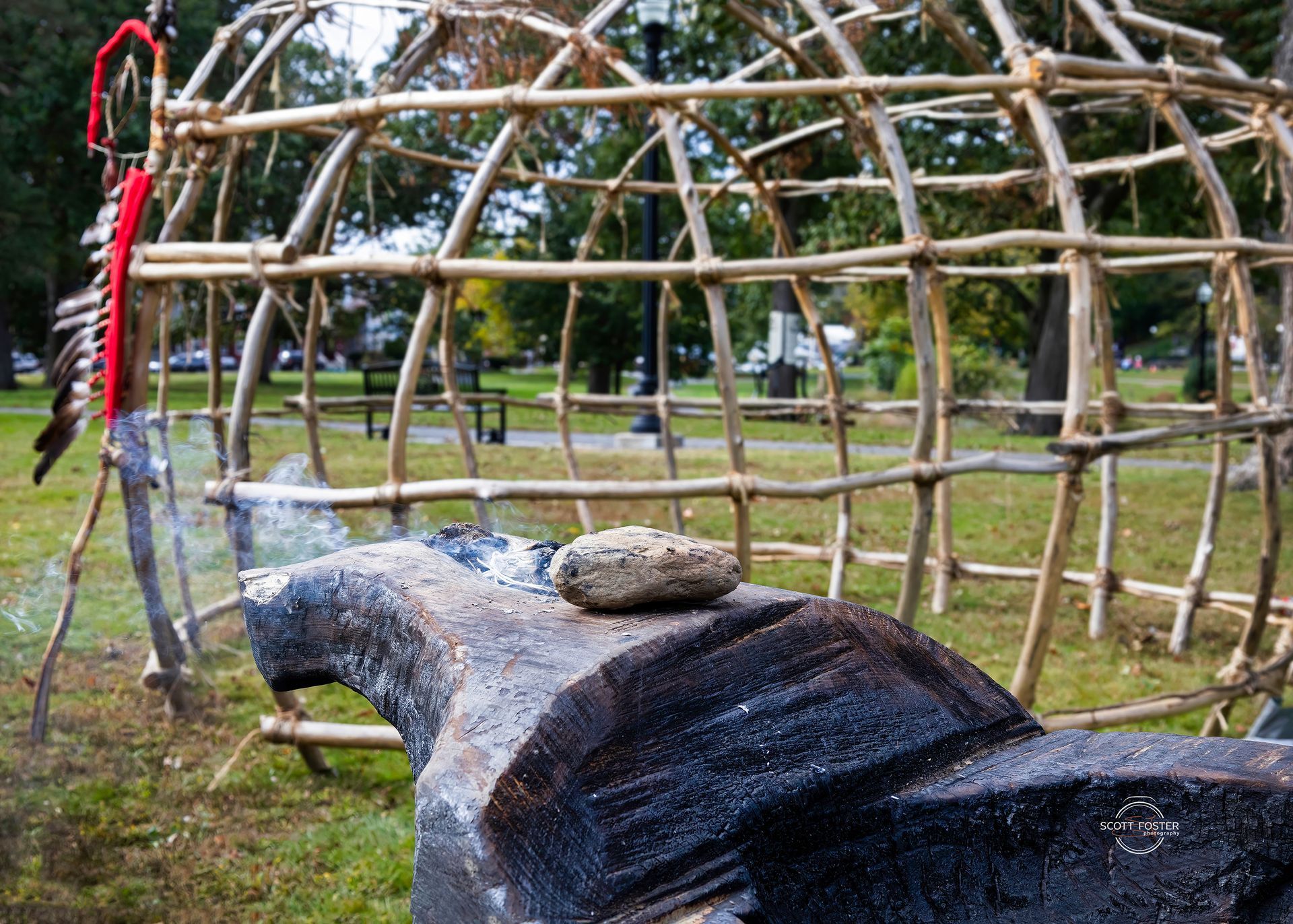 Wooden frame of a Native American dwelling with a stone on a dark, carved structure; smoke visible.