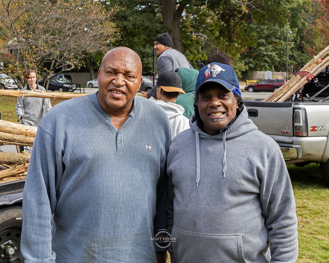Two men posing outdoors with a truck in the background, surrounded by logs and other people.