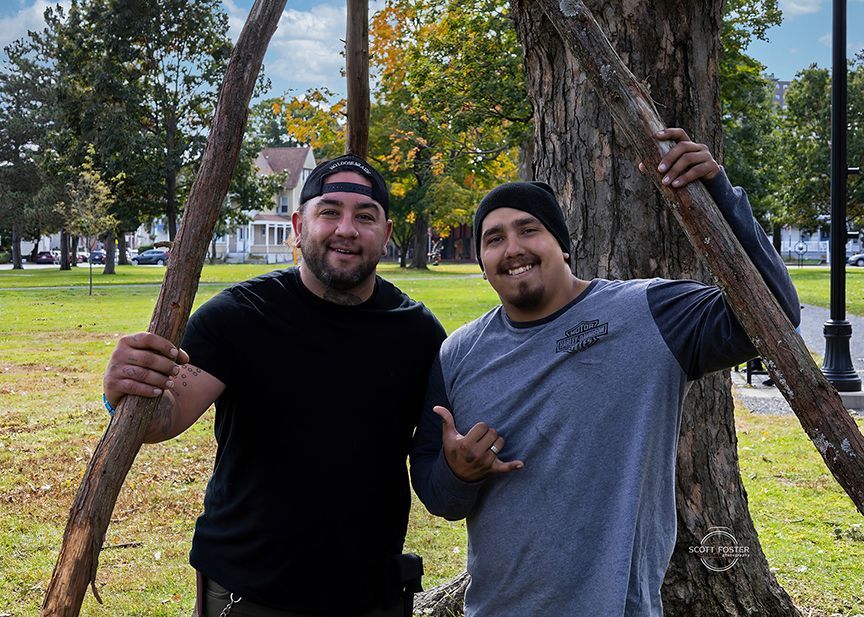 Two men smiling, holding tree supports in a park. One in a black shirt and hat, the other in a gray shirt and beanie.