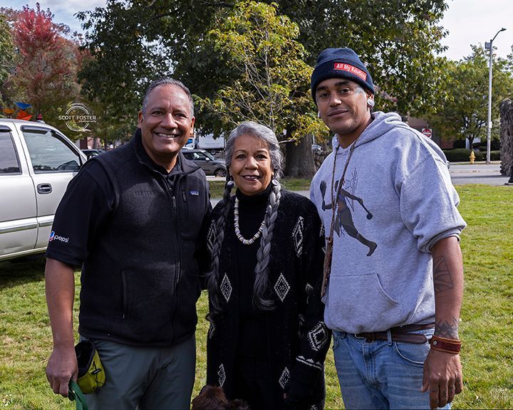Three people smiling outside. Man in black, woman in black sweater, man in gray hoodie. Green grass, trees.