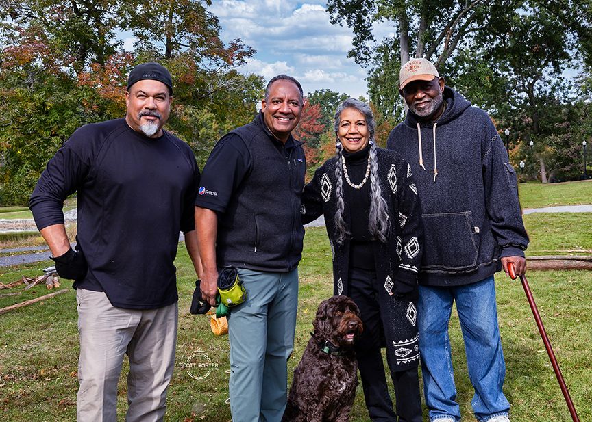 Four people, one with a dog, pose outdoors. Green grass and trees in background.