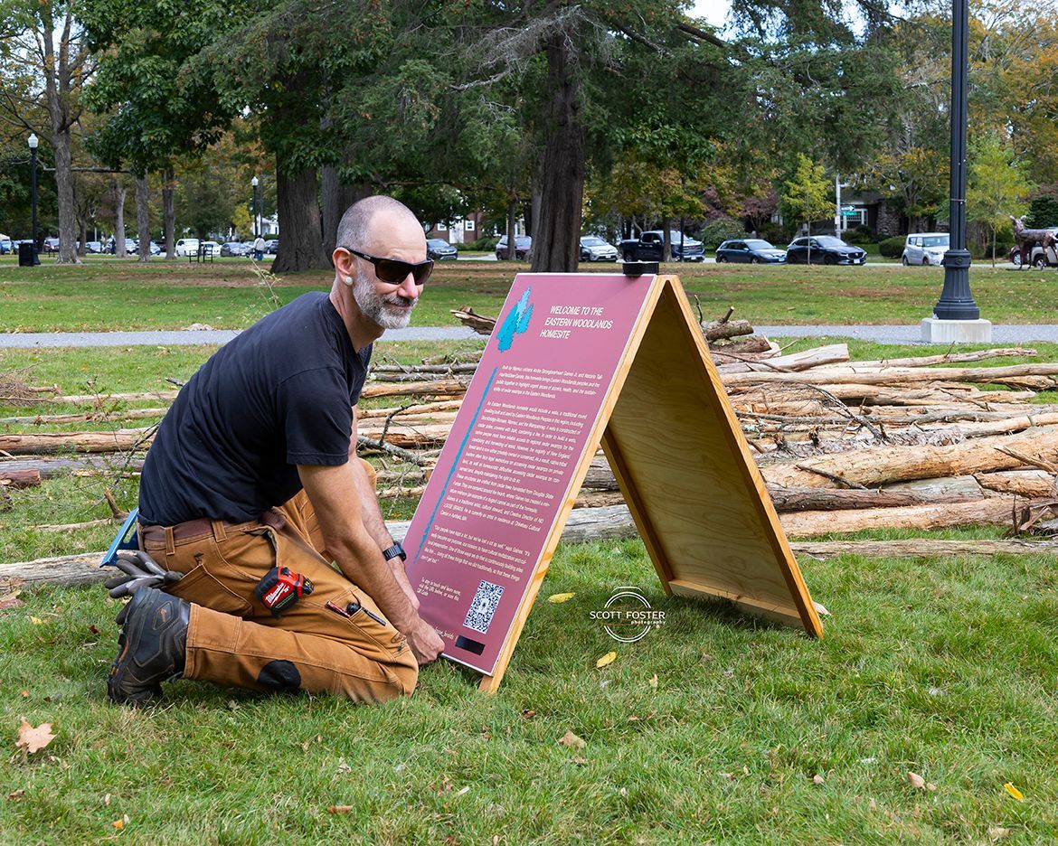 Man installing an A-frame sign on grass near stacked logs. He wears a dark shirt and work pants.