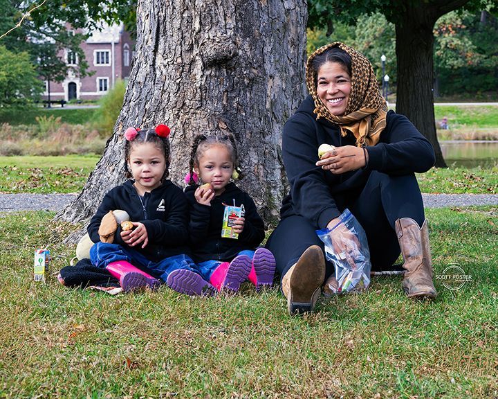Woman and two children sit under a tree, enjoying snacks in a park.