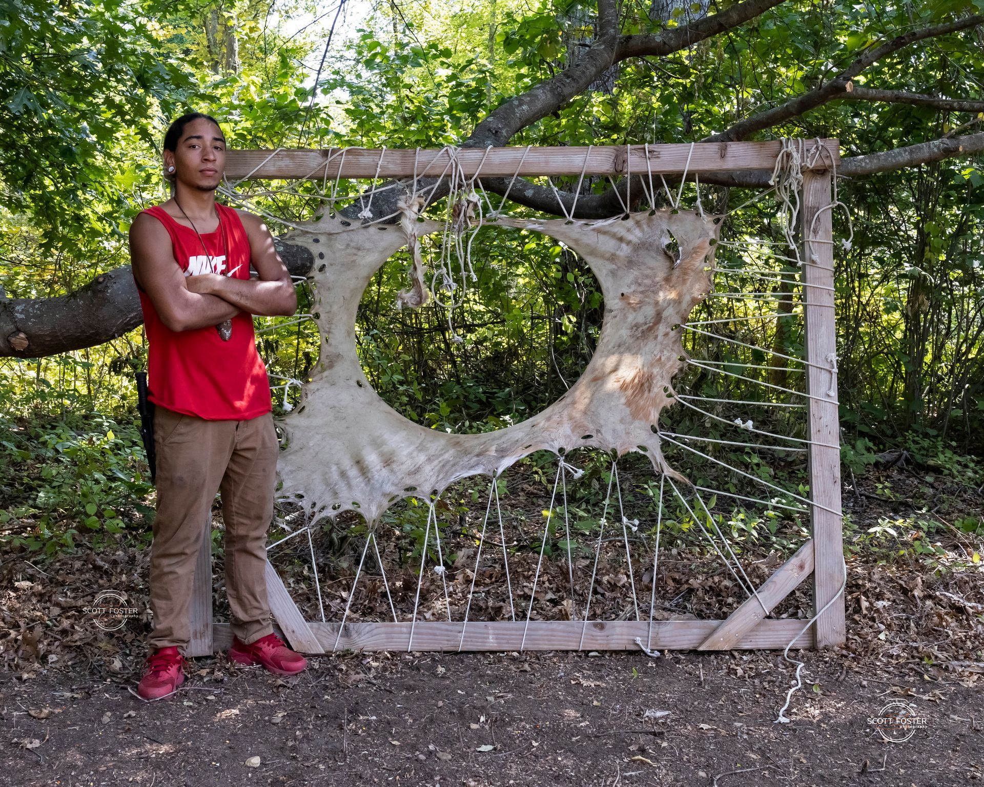 Person stands by a wooden frame with stretched hide. Person wears red shirt, khaki pants, outdoors.