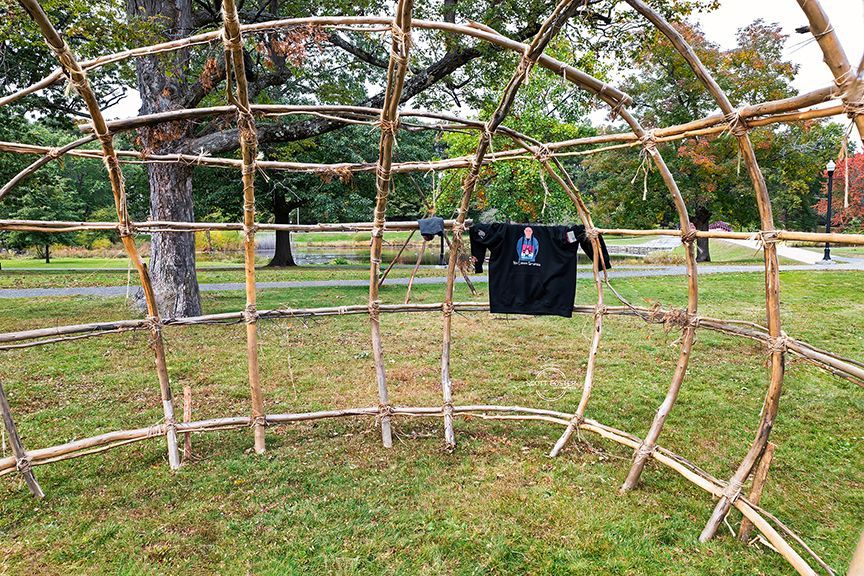 Wooden frame structure on grass, with a shirt hanging inside. Trees and green space in the background.