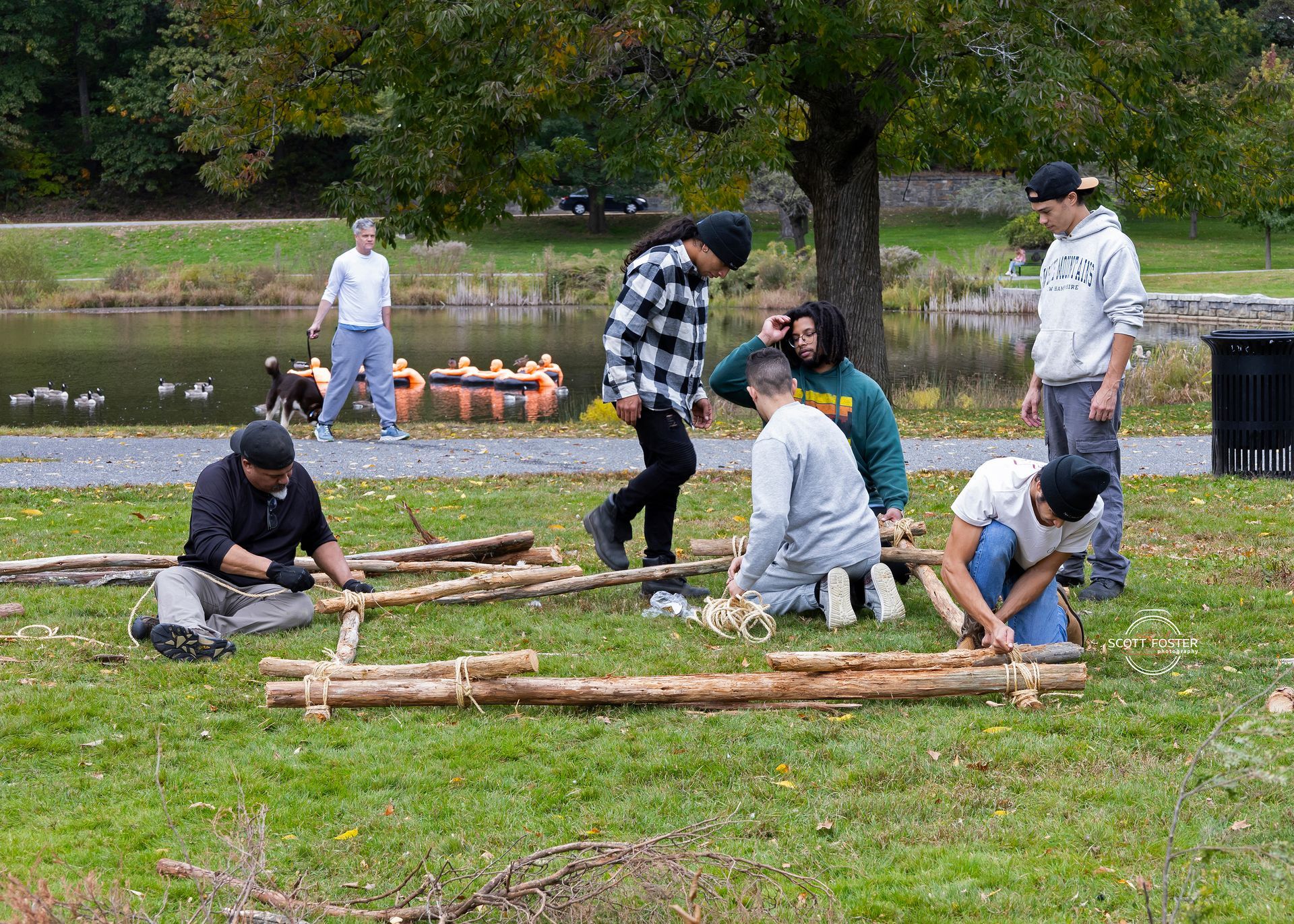 Group of people building with logs in a park, near a pond with ducks, on a sunny day.