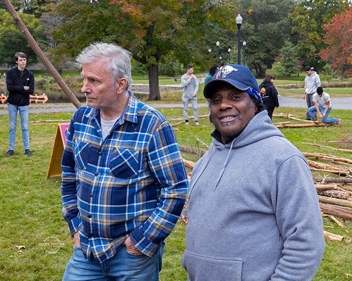 Two men in a park: one in flannel, the other in a hoodie. People work with wood in the background.