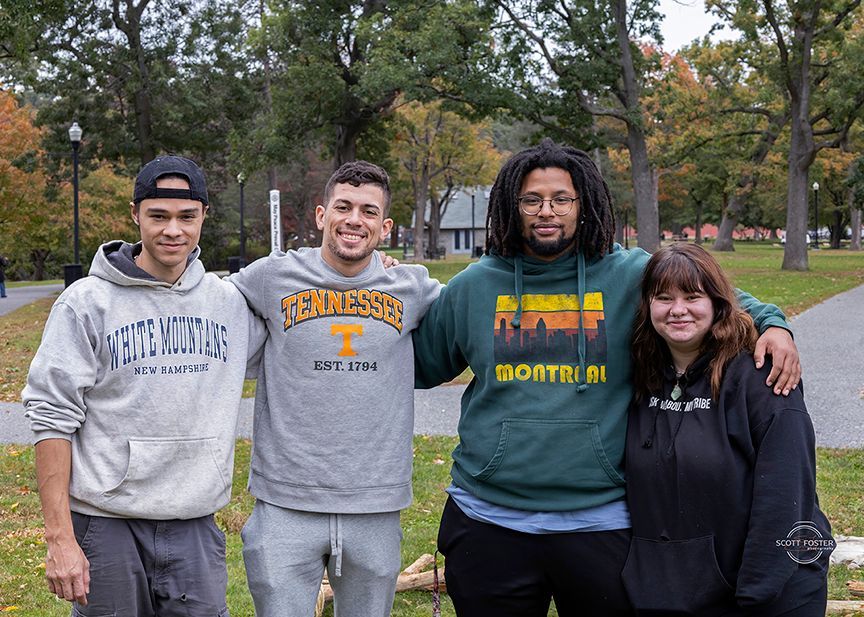 Four people standing close together outdoors, smiling. Two men have their arms around two others. 