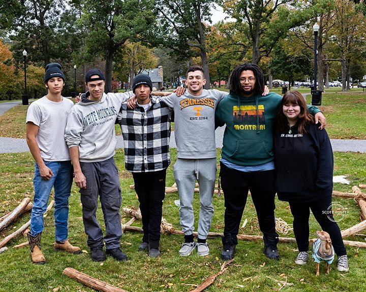 Group of six people with arms around each other stand in a park next to wooden logs; green trees.