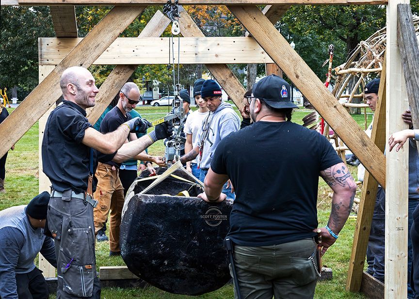 People lifting a large, dark object using a wooden hoist in an outdoor setting.