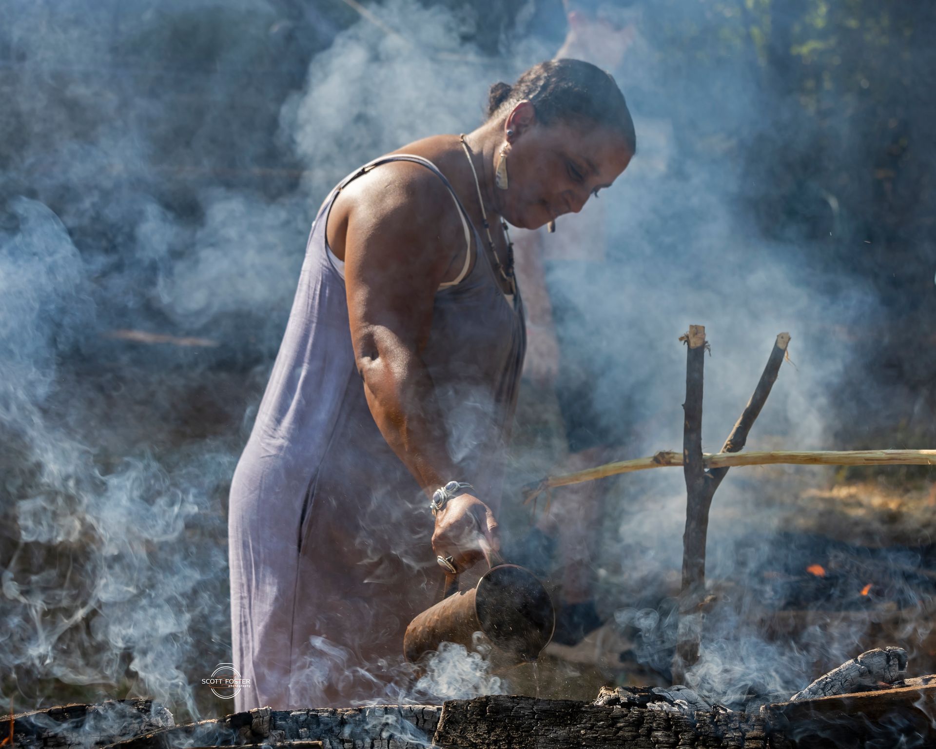 Woman tending a fire, stirring with a wooden implement. Smoke surrounds her in a sunny outdoor setting.