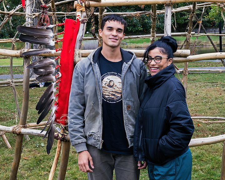A man and woman stand by a wooden structure adorned with feathers, smiling. Outdoor setting.
