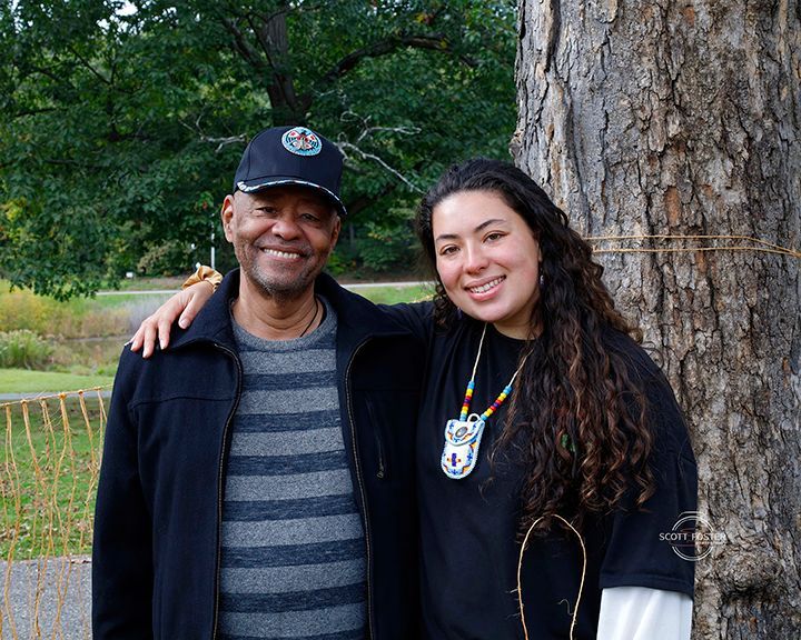Two people stand by a tree outdoors. One man in a hat and jacket has his arm around the woman. Both are smiling.