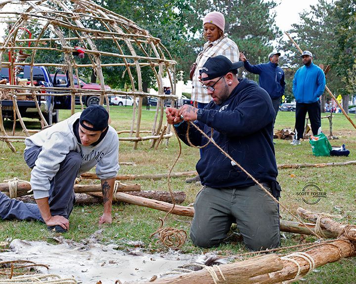 People constructing a wooden structure outdoors. One man kneels, tying cord; another squats nearby. Others stand and observe.